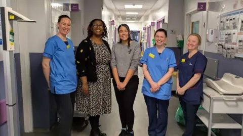 Birmingham Children's Hospital Jasmine Page is pictured smiling on a hospital ward, she is standing in between four nurses. She wears a grey jumper and black trousers with black trainers. Three nurses are wearing blue nurses uniform with their hair away from their faces while another nurse wears a black and white floral dress, black and yellow cardigan and glasses. 