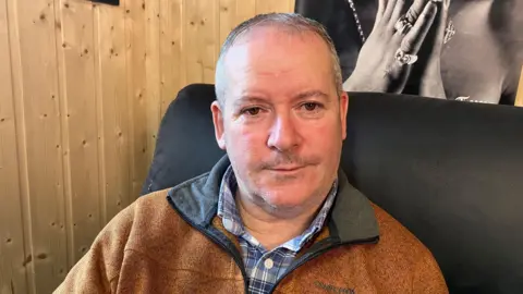 Sean Bruen sits on a black chair, he has grey hair, brown eyes and is dressed in a blue checked shirt with a brown coat over it. There is a poster on a wooden wall behind him.