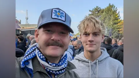 Craig Finbow A selfie of Craig and his son Oscar outside the ground. here are crowds of people behind them, mostly men. Craig wears the blue and grey Ipswich Town scarf and a grey-blue baseball cap with a flower on the front. Oscar wears a grey hoodie.