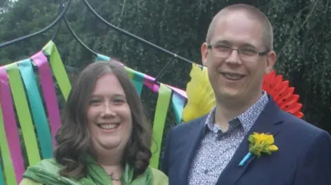 Vicky wears a green dress and has shoulder-length brown hair. Her husband wears a blue suit jacket with a yellow flower in its lapel, a patterned blue and white shirt, and has closely-cropped hair.