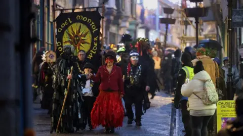 Penzance Council A group of people walking down the street holding a sign which reads Goel Montol, one of the people is wearing a red mask, hat and red jacket and dress, others around them are wearing costumes and masks.