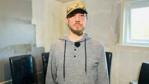 Gary Bennett, a man wearing a baseball cap and grey hooded top, in front of mouldy furniture in his council flat. 