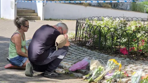 Stuart Woodward/BBC A man and a woman kneeling by floral tributes left in the park. The man is visibly emotional with his head buried in his arms. There is police tape in the background.