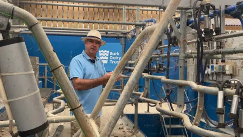 Major beaumont gestures at the dairy farming equipment, wearing a blue polo top and straw hat. The equipment is covered in cobwebs and dust, with a blue and white "Sark Dairy Trust" sign in the background.