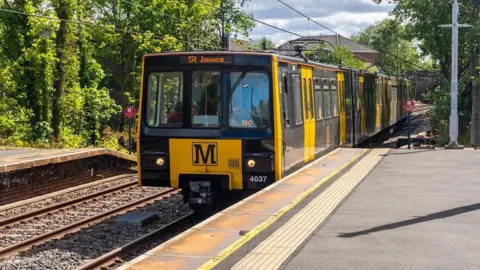 A black and yellow Metro pulling into a station with 'St James' written on the front. Two concrete platforms sit on either side of the track.