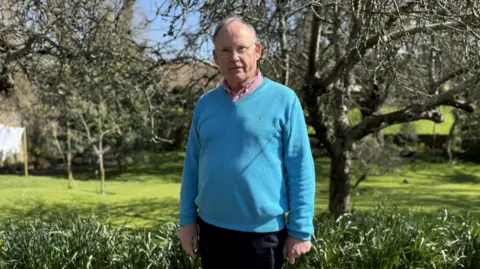 A man in his 70s with short grey hair. He is wearing glasses which have no frame, a bright blue jumper. You can see the collar of his pink shirt on the neck line.