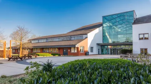 A large office building with a middle section which has a tall glass frontage. The section to the left has a sloping roof on two levels, and to the right there is a standard two-storey building. There are benches on the pavement area in front with plants in a bed.