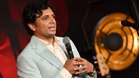 Warner Bros M. Night Shyamalan wears a white shirt and a blue pinstripe blazer