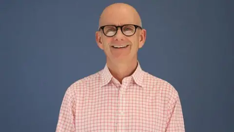 NHS England South West A man smiling at the camera. Here is wearing a pink chequered top and glasses. The background is blue. 