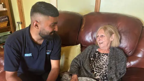 A woman sat on a brown leather sofa talking to her male carer