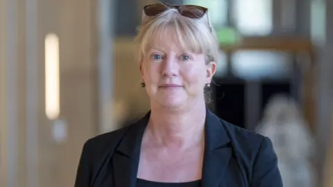 PA Media Head and shoulders shot of Shona Robison, a lady with tied back blonde hair, walking along a brightly lit corridor in the Scottish Parliament building in Edinburgh. She is looking directly at the camera and is slightly smiling. She is wearing a black top, matching jacket and has a pair of sunglasses balancing on her head.