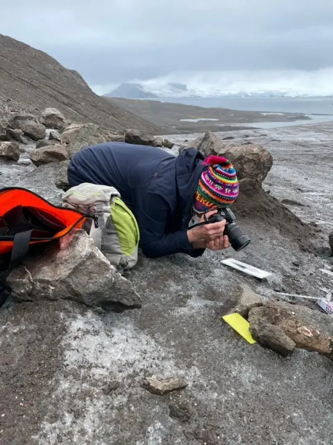 Dariusz Puczko A person wearing a colourful woollen hat and outdoor clothes kneels on icy surface pointing a large camera at some rocks, with items around it to give a sense of marking a position.