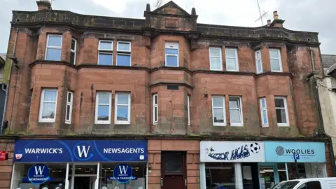 BBC news A large sandstone building with multiple windows and a shop on either side of a burgundy door on the ground floor 