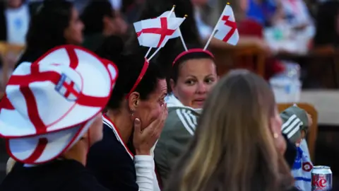 PA Media A group of women at a watch party for an England football match in Cheltenham. They are wearing England merch and one woman has her head in her hands reacting to a goal from the opposition.
