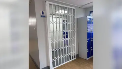 A white gate with ornate bars across the entrance to toilets at Cottingham High School. Blue lockers can be seen to the right.