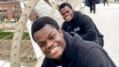 James and John Makanjuola James and John Makanjuola are outside on a bridge, cleaning. They are both looking at the camera and smiling, with one behind the other and wearing matching black hoodies and tracksuit trousers.