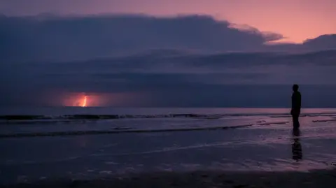 Aivaras Sakurovas Lightning in the distance over the sea on Crosby beach. A figure on the sand is shown in shadow on the right. 