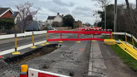 Tony Fisher/BBC A large hole which has been dug in the road. There are plastic safety railings around it. There are more railings in the background. On the right is a hedge and on the left there is a row of houses.