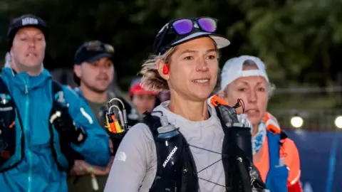 Rich Gill Stephanie Case is in the foreground of the photo. She wears a grey long-sleeved running top with a black hydration vest over it. She has a peaked black and grey cap on with sunglasses above the visor. One red earbud can be seen in her right ear. She is looking towards the right of the photo. A female runner is to her right and behind, and two male runners are behind her to the left. All three are slightly out of focus.