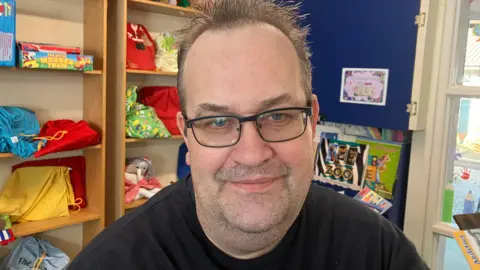 Paul Scocroft has brown hair and wears glasses. He is wearing a black jumper. He is behind shelves with books and toys.