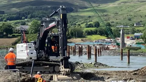 Construction work on the Narrow Water Bridge project, with large black digger in front and men with amber tops.