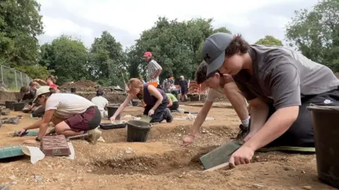 A group of people, some young students, others retired volunteers work on their hands and knees removing soil from a graveyard at the dig site.