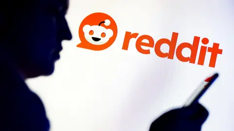 In this photo illustration, a silhouetted individual is seen holding a mobile phone with a Reddit logo displayed in the background
