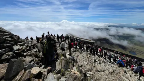 Olly Outdoors This image shows the summit of Snowdon, with queues of people waiting to reach the top.