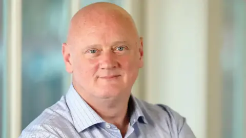 FICO Campbell Scott looking directly at the camera. He is bald. He has blue eyes and is wearing a light blue shirt with dark checks on it.