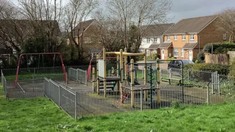 A view of a play park on Blackthorn Drive in Ivyrbridge. There's a silver fence around the park area which has a variety of equipment. The area is surrounded by grass with houses to the right.