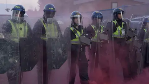 Police officers stood with shields surrounded by pink dust amid clashes with protesters