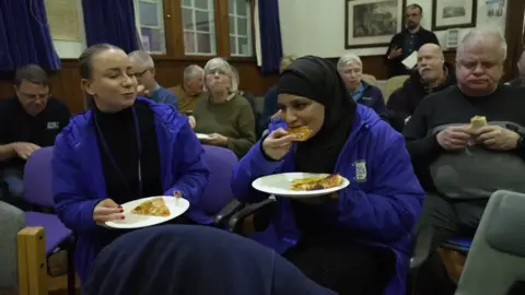 Two women laugh together as they eat pizza in a room full of people