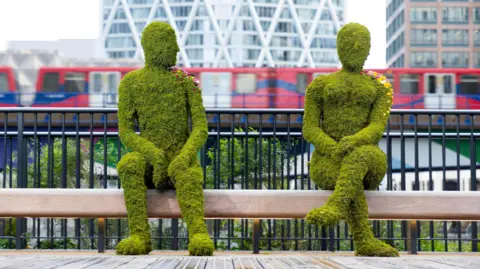 Two figures, larger than lifesize, one male and one female, made out of moss. They are 'sitting' on a bench as a London train passes in the background