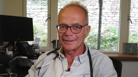 MP Dr Simon Opher sits in the chair in his surgery smiling at the camera. He is wearing a pale shirt and has a stethoscope around his neck, and is also wearing black-framed glasses