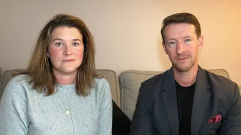 Vicky and Douglas sit next to each other on a sofa and look at the camera. Vicky is wearing a grey jumper with a necklace and has light brown hair which comes down to just below her shoulders. Douglas has a dark grey jacket on with a spotted red pocket square and has a black t-shirt underneath. He has short dark hair which has been brushed to the side.