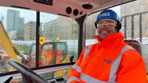 A woman sits in a dumper truck cab. She has hi-viz on and a blue helmet. She is smiling.