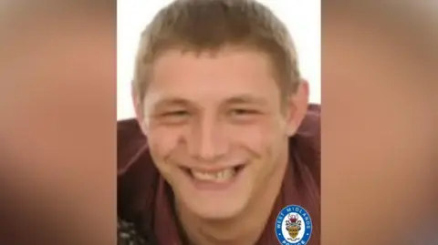A man with short light brown hair and a maroon top. He is smiling. The background is blurred and there is a West Midlands Police logo at the bottom.
