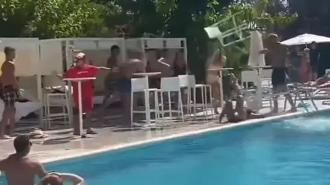 BBC A group stood by a pool as a white chair flies through the air towards a man's head