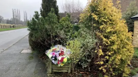 Steve Jones/BBC Shop-bought flowers placed in a flower box in a front garden. Conifers surround the box. A pavement and a road is visible to the left of the picture.