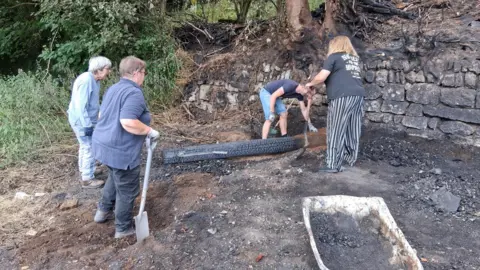 Shelley Porter The remains of the shelter. The image shows scorched ground and burnt items in front of a stone wall and trees. Four people are helping to clean debris.