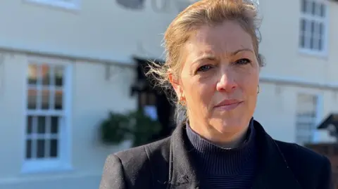 Polly Durant stands in front of her pub. She has light brown hair that is tied up behind her head. She wears a black coat with a blue jumper underneath.