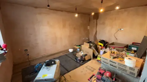 A room has been freshly plastered and has new light fittings in the ceiling. There are several construction tools and boxes around the room.