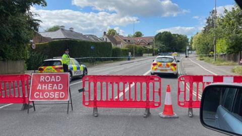 A red road closure sign on Woodstock Road, with red barriers closing off the road in the foreground and a police tape cordon, three police cars and a police officer in the background, further along the road