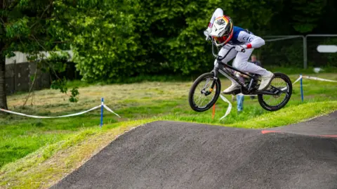 Pete Scullion A rider dressed in white on a black bike takes off on a bump on a track