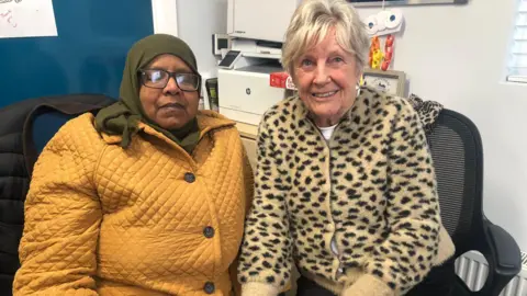Hala Ahmed is on the left of the image wearing a head scarf, yellow caot and glasses. To her right is Jeanette Warke wearing a yellow coat on which there are blackspots. She has blonde hair. Both ladies are sitting side by side.