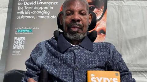Syd sitting in a wheelchair with a dark polo shirt on, short beard and hair, smiling at the camera. There is a poster behind him and he is outside by a white tent.