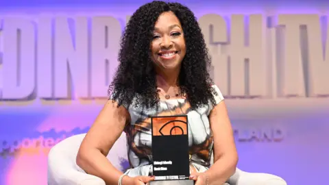 Edinburgh TV festival Shonda Rhimes, with a bright sile and dark curls, holds her award sitting on set at her TV festival show. The background is pale purple and she wears a patterned white, orange and black dress while sitting on a white chair.