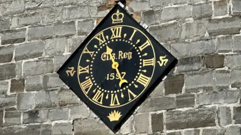 BBC Sitting on a stone church wall is a diamond-shaped clock, with a golden face and hand. 