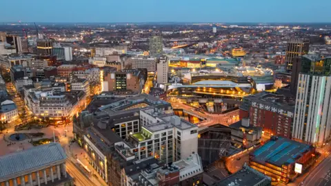 A view of Birmingham city centre taken from the sky. It is early evening as the buildings are visible but the lights and street lights are on. The Rotunda can be seen in the distance in the middle of the shot.