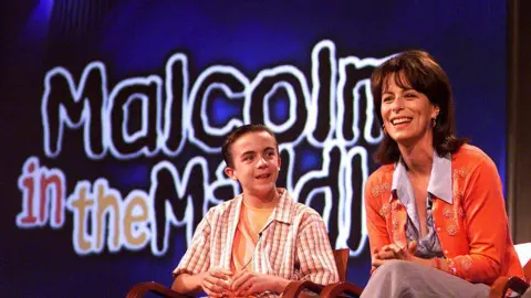 Getty Images Jane Kaczmarek, who has brown shoulder length hair and is wearing an orange cardigan, sitting next to Frankie Muniz, who is wearing an orange T-shirt underneath a white and brown checked shirt, at a press event for Malcolm in the Middle.